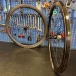 Due cerchi WS-ROAD-Revolution-CarbonSpokes-Hookless-1162gr appoggiati su un tavolo da lavoro scuro e parete con attrezzi